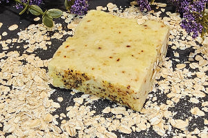 Bar of soap with visible ingredients on a black surface with oats and lavender flowers.